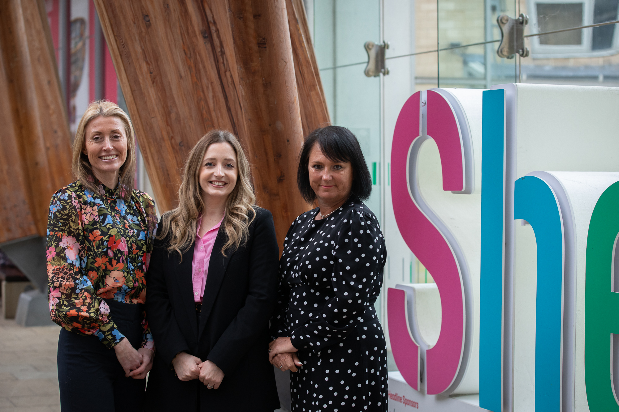 Three people standing indoors next to large wooden beams and a colorful sign that partially reads “She” in bold pink and blue letters. The setting appears modern with glass panels and natural light coming through.