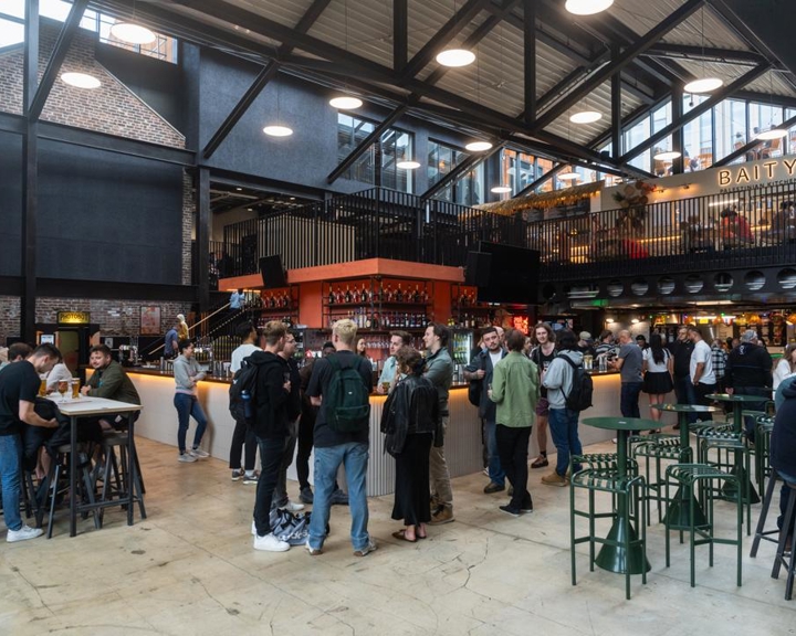Interior of a modern food hall with high ceilings, exposed beams, and industrial-style lighting. The space is busy with groups of people standing and sitting at tables, socializing and enjoying drinks. At the center is a large bar with an orange canopy, surrounded by bar stools. In the background, there are multiple food stalls and a mezzanine level with additional seating. A sign reading “BAITY” is visible on the upper level.
